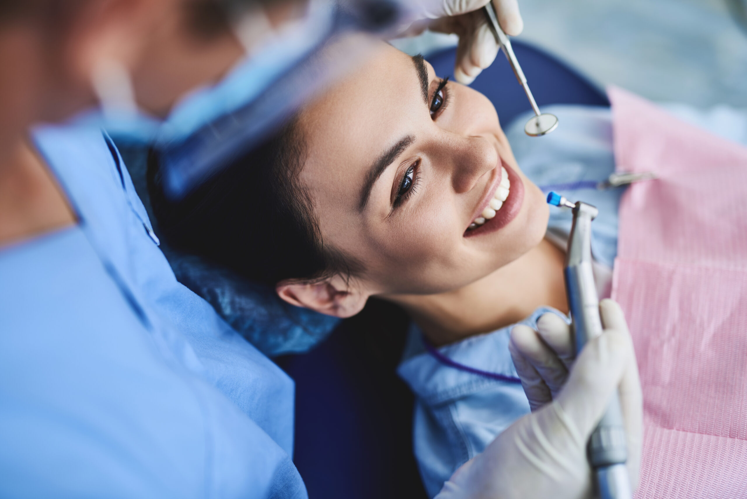 Close up portrait of charming woman sitting in dental chair while stomatologist holding polisher and mirror. Girl is smiling